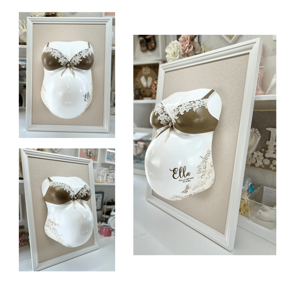Bilderrahmen in Sandbeige mit Babybauch-Gipsabdruck, veredelt mit braunem Spitzen-BH, braun-cremefarbenen Ornamenten, Schmetterling an der Seite sowie dem Babynamen und den Geburtsdaten – professioneller Veredelungsservice für Schwangerschaftsabdrücke.
 - Atelier Body - pArts