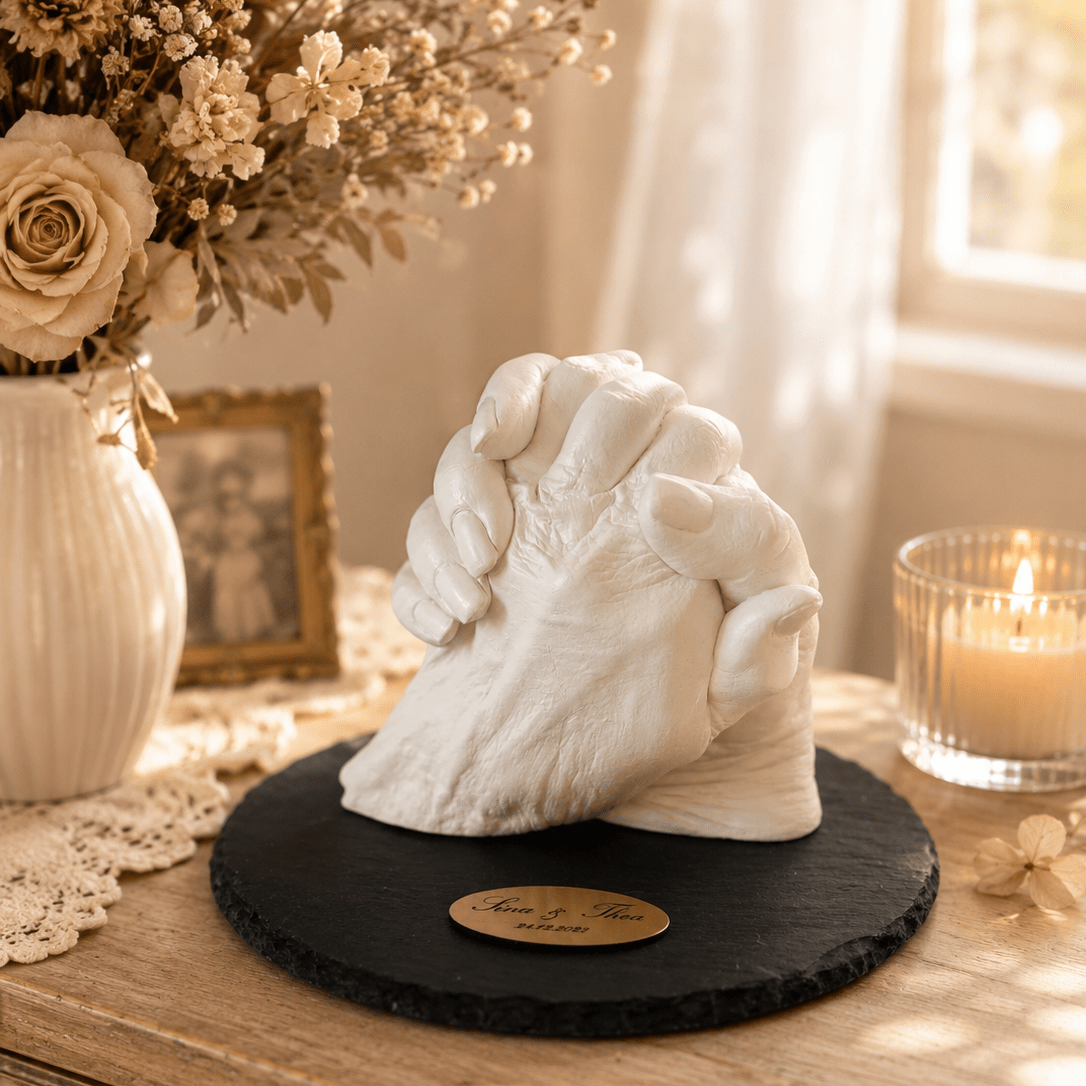 Das Bild zeigt einen detailreichen 3D-Händeabdruck von zwei Personen auf einer eleganten Schieferplatte mit personalisierter Gravur der Namen und des Datums der Abformung. Der handgefertigte Gipsabdruck symbolisiert Verbundenheit, Familie und gemeinsame Erinnerungen und dient als zeitloses Erinnerungsstück für Generationen.- Atelier Body - pArts