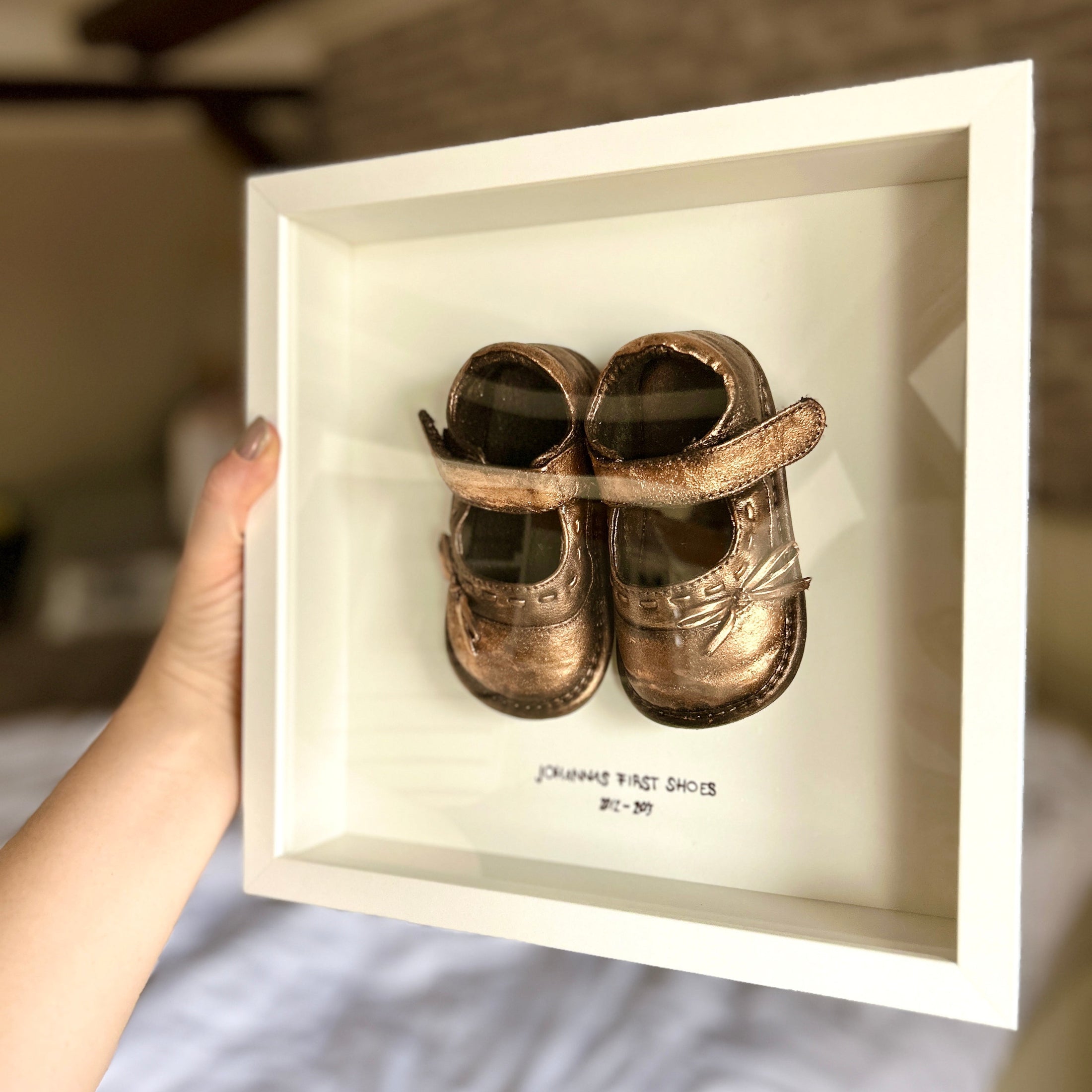 Weißer Erinnerungsrahmen 25x25 cm mit den ersten Babyschuhen, handveredelt in Bronze personalisiert mit Namen des Kindes – liebevolles Babyandenken aus dem Atelier Julia Schulze