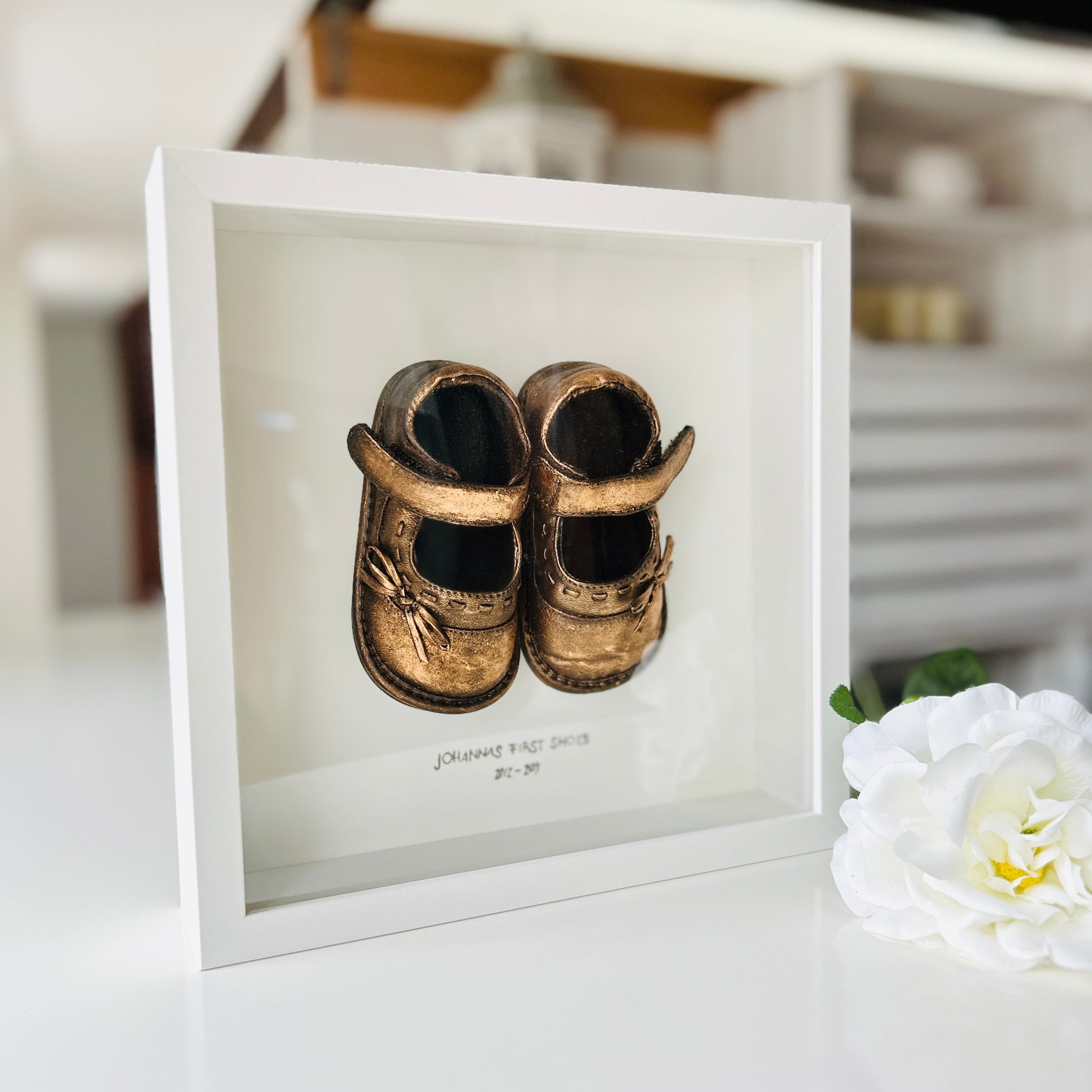 Weißer Bilderrahmen mit echten Babyschuhen, veredelt in Edelmetallfarbe, individuell beschriftet mit Namen – exklusives Baby-Geschenk aus Handarbeit.