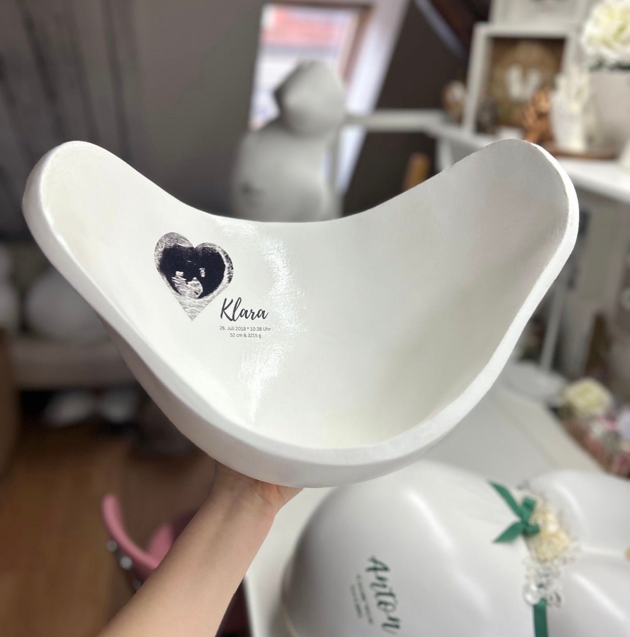 Das Bild zeigt eine elegante weiße Belly Bowl aus einem Babybauch Gipsabdruck mit schlichtem schwarz-weißem Design. Im Inneren der handgefertigten Bauchschale befindet sich ein kleines Herz mit Ultraschallbild sowie der Name des Kindes und die Geburtsdaten. Die Belly Bowl wurde professionell verspachtelt, geschliffen und lackiert und dient als stilvolle Erinnerung an Schwangerschaft und Geburt sowie als dekorative Schale für kleine Erinnerungsstücke, Babysachen. Atelier Body - pArts