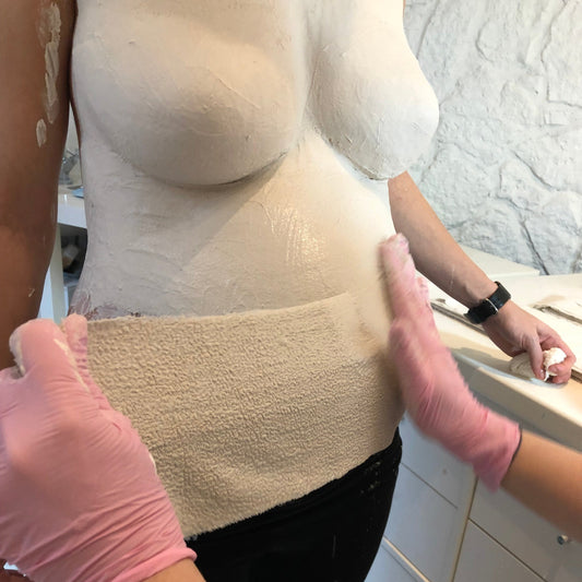 Das Bild zeigt die Abformung eines Babybauchs mit Gipsbinden während der Schwangerschaft. Die werdende Mama steht entspannt, während der Bauch sorgfältig mit mehreren Gipsbinden modelliert wird, um die besondere Form der Schwangerschaft als bleibende Erinnerung festzuhalten. DIY-Babybauchabdruck zur späteren Veredelung oder Gestaltung im Atelier Body-pArts von Julia Schulze.