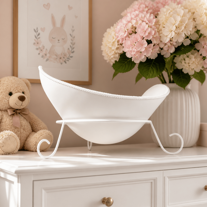 Das Bild zeigt eine schlichte und elegante weiße Belly Bowl aus einem Babybauch Gipsabdruck mit matter Lackierung und feiner weißer Posamentenborte. Die handgefertigte Bauchschale steht auf einem weißen Metallsockel mit drei Füßen auf einer stilvoll dekorierten weißen Kommode im Kinderzimmer. Blumen wie Hyazinthen, ein kleiner Teddybär und helle Einrichtungselemente unterstreichen die ruhige und hochwertige Atmosphäre. Die Belly Bowl integriert sich harmonisch als modernes Wohnaccessoire.